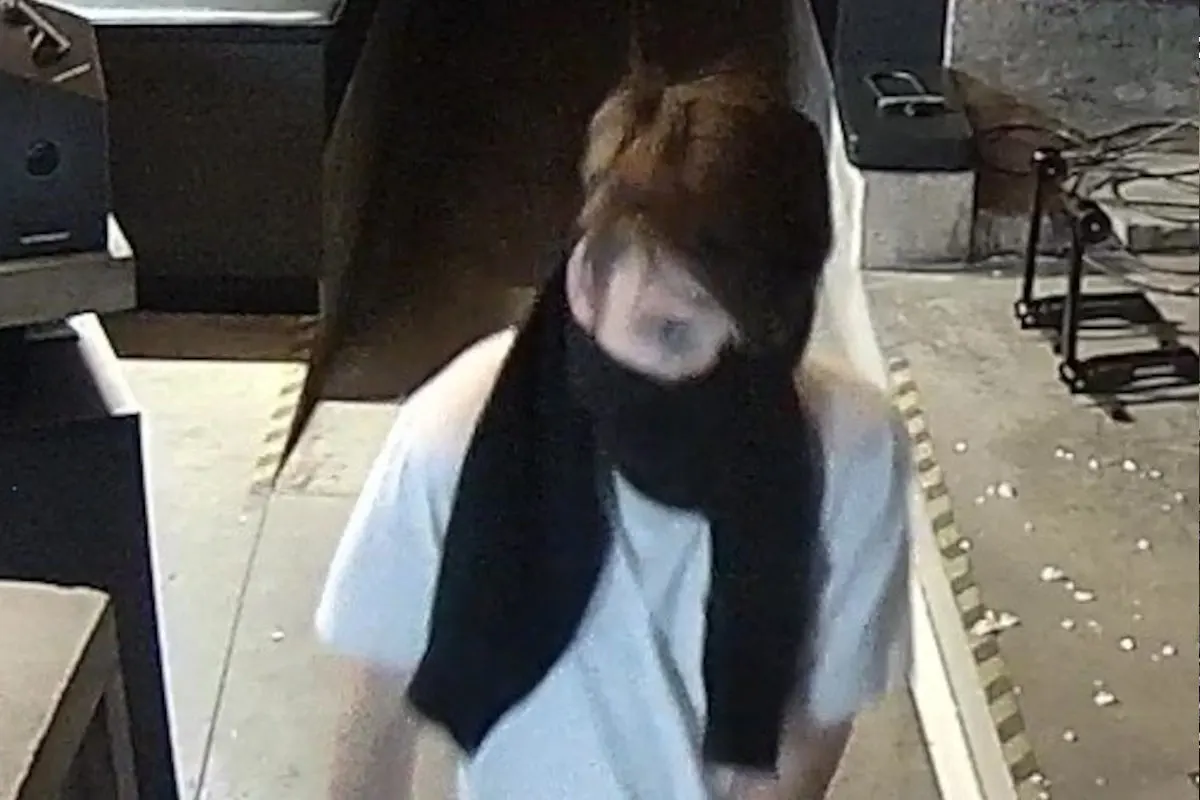 Eine Person trägt einen schwarzen Schal, der das Gesicht verdeckt, in einem überdachten, industriellen Umfeld. Der Boden ist teilweise mit Schmutz bedeckt.