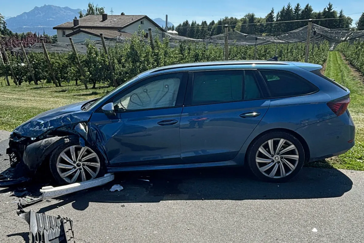 Ein blauer Kombi steht mit beschädigter Front auf einer Landstraße vor einem Haus. Im Hintergrund sind Berge und Bäume zu sehen.