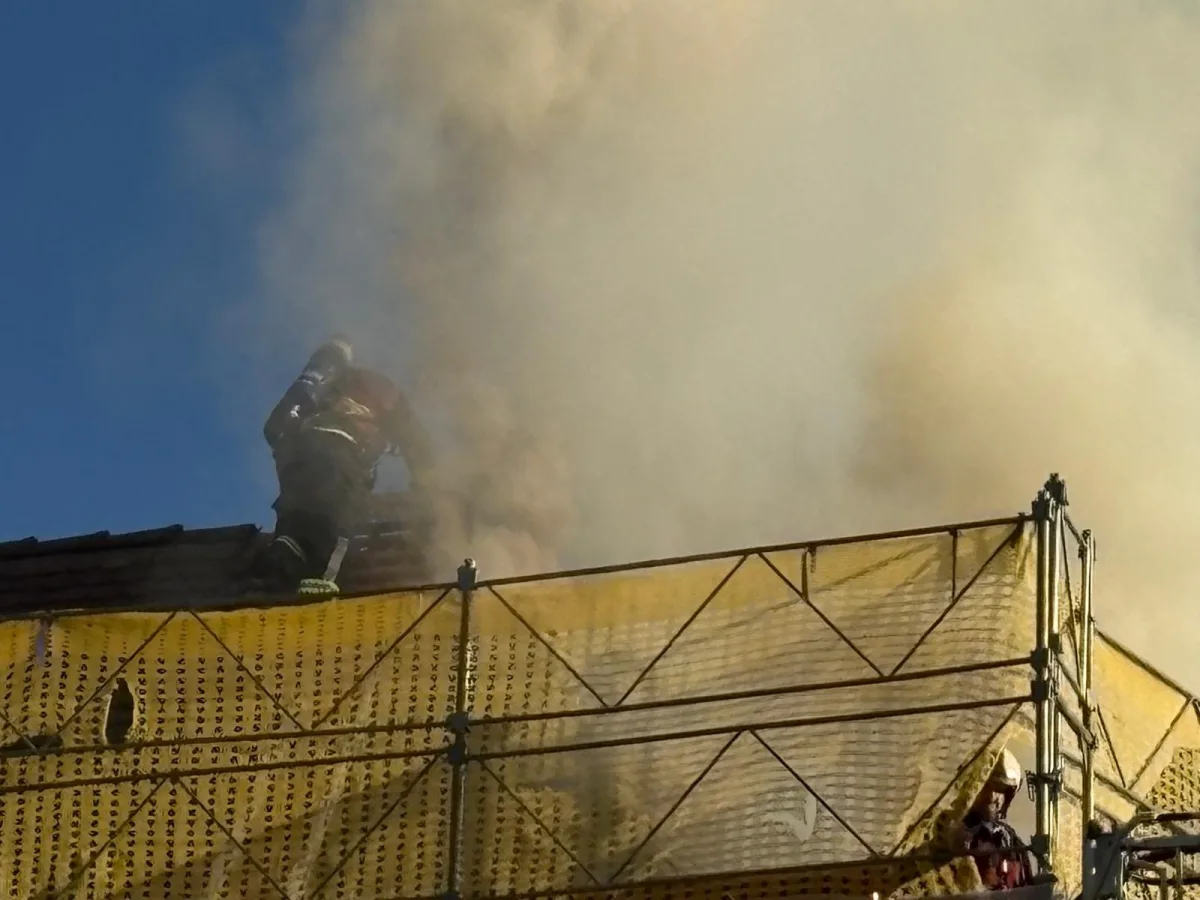 Ein Feuerwehrmann arbeitet auf dem Dach eines Gebäudes, das stark qualmt. Er trägt Atemschutzgeräte, während Rauch um ihn herum aufsteigt.