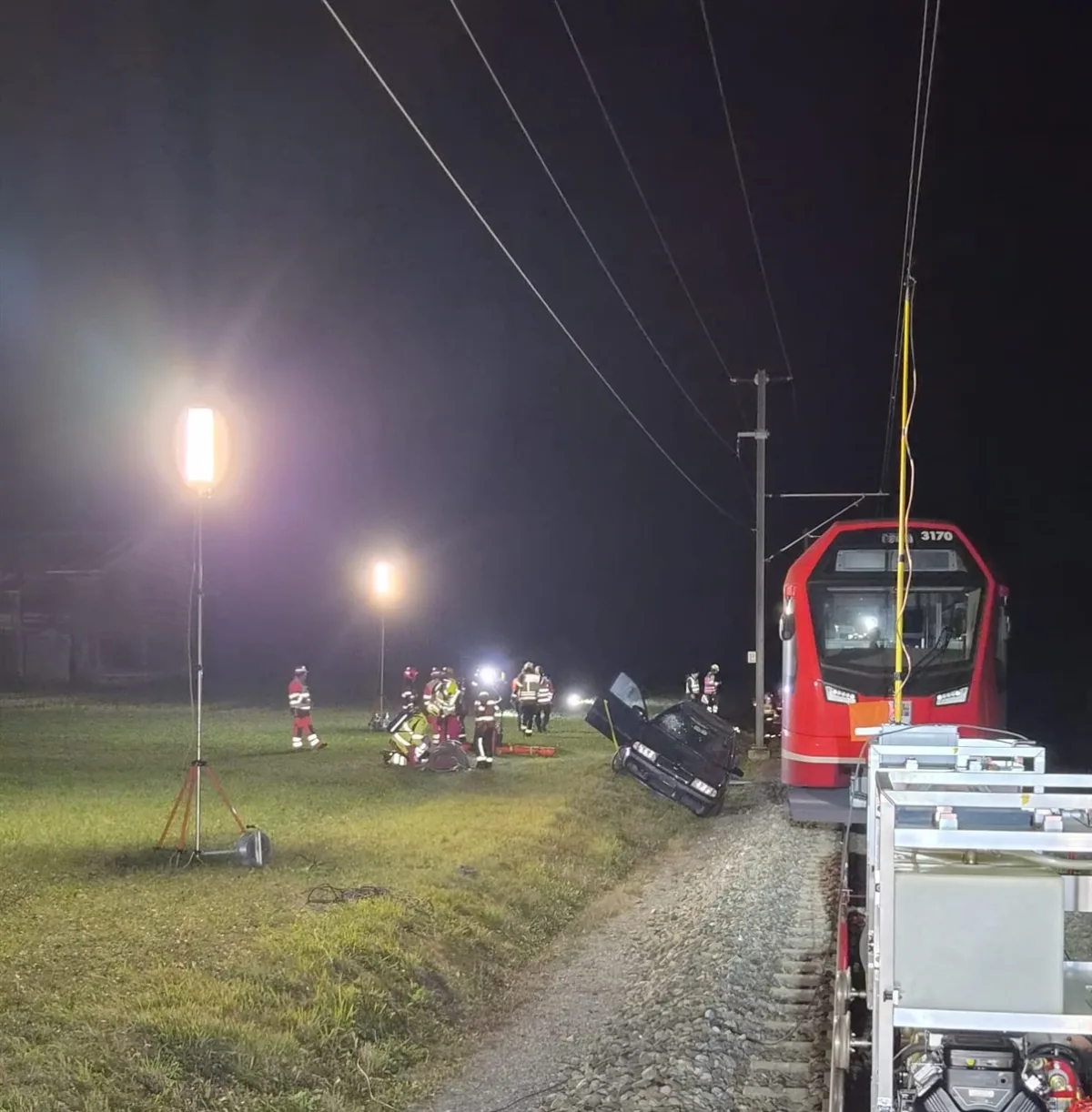 Ein roter Zug steht neben einem verunfallten Auto, Rettungskräfte arbeiten vor Ort. Einsatzlicht beleuchtet die Szene.