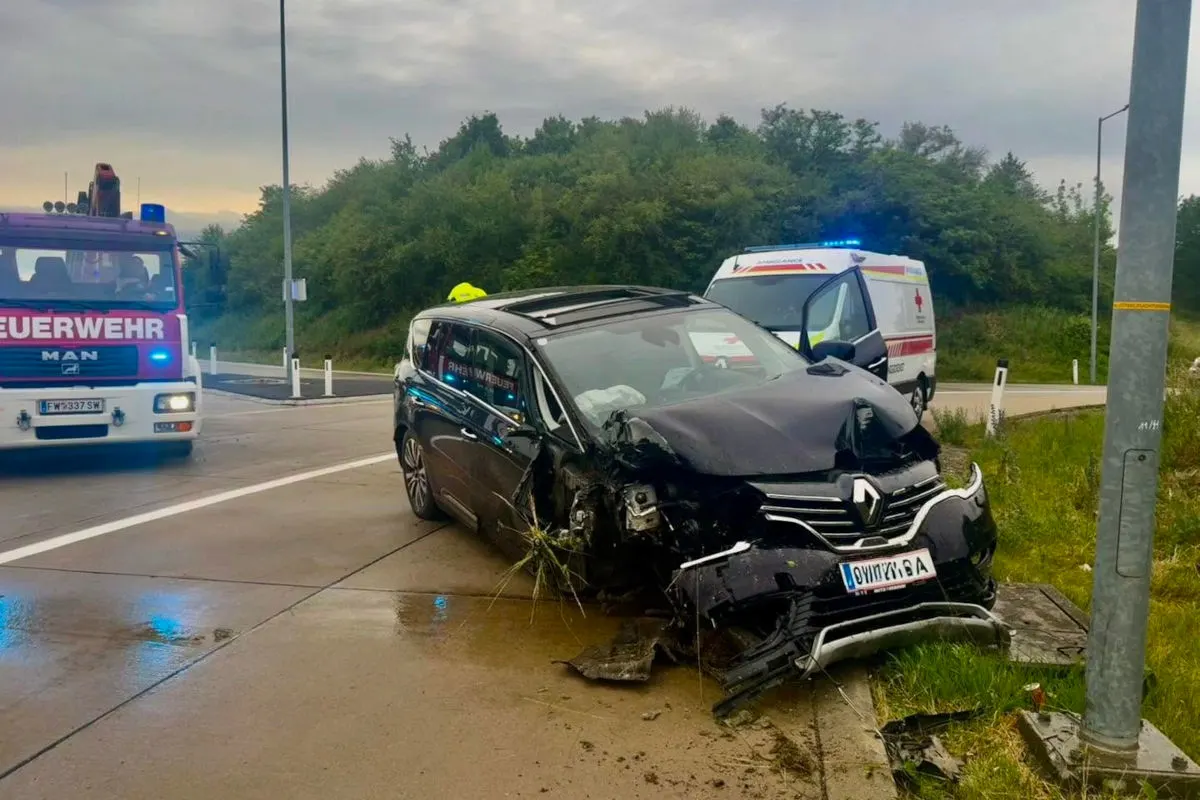 Ein stark beschädigter schwarzer Renault steht neben einem Lichtmast auf der Straße. Ein Feuerwehrauto und ein Krankenwagen sind im Hintergrund zu sehen.