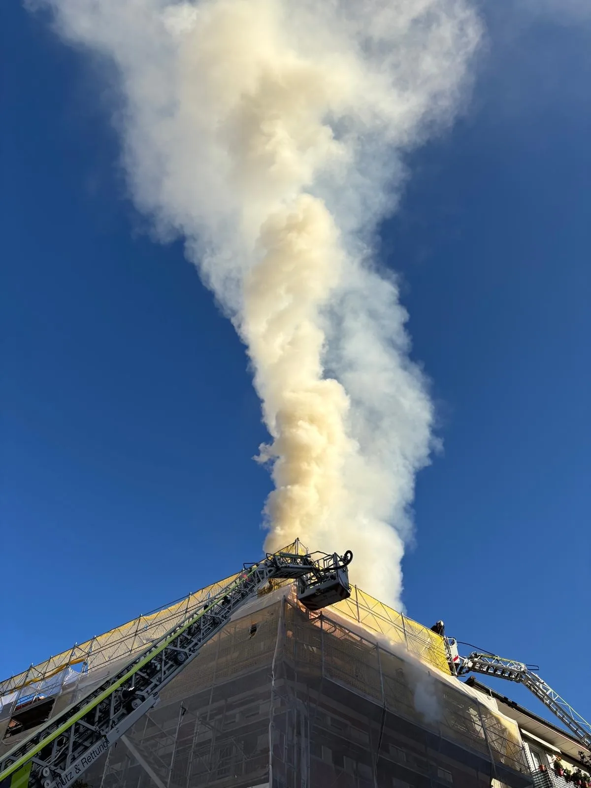 Ein Baugerüst umgibt ein Gebäude, dessen Dach in Flammen steht, mit starkem Rauch, der in den Himmel steigt.