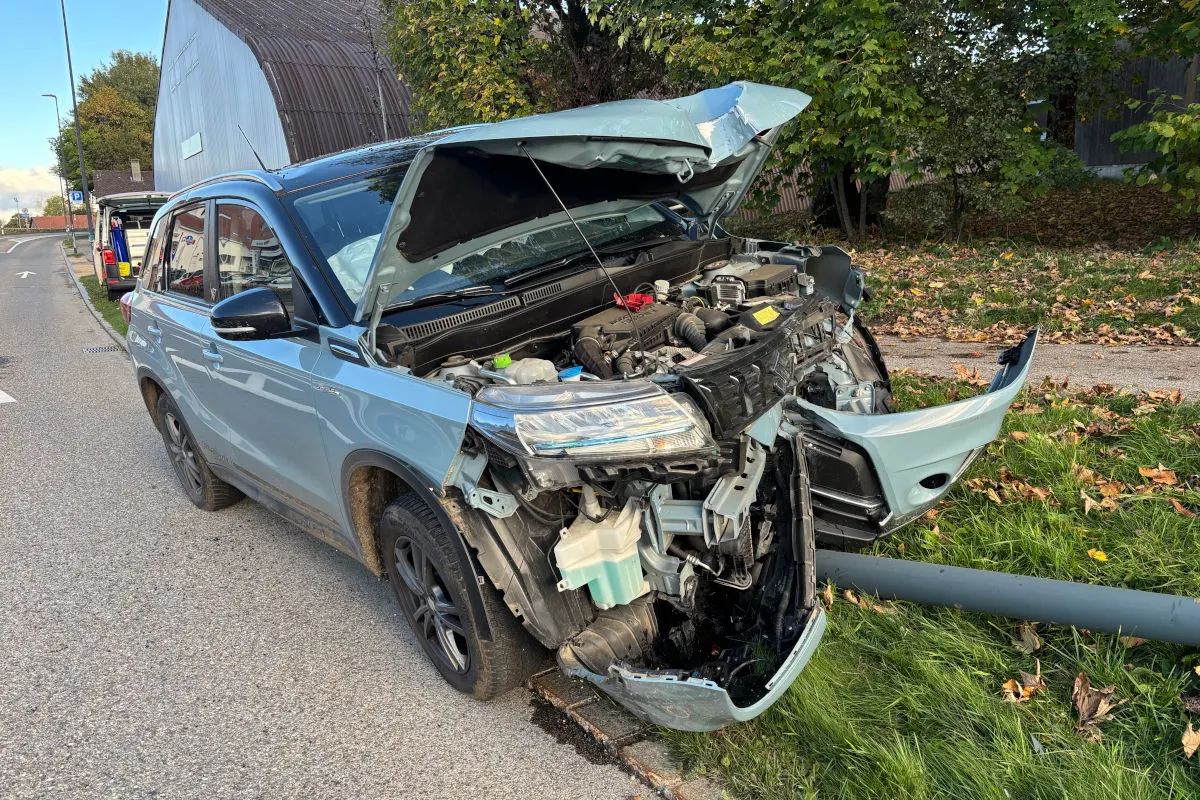 Ein hellblaues Auto mit stark beschädigtem Frontbereich steht auf einer Straße neben einem gefallenen Baumstamm. Die Motorhaube ist geöffnet.