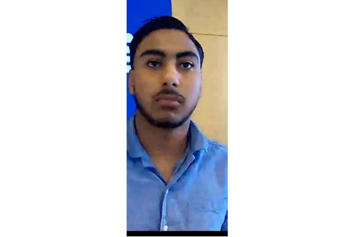 Ein Mann mit dunklem Haar und Bartansatz trägt ein blaues Hemd. Er steht vor einem blauen Hintergrund.
