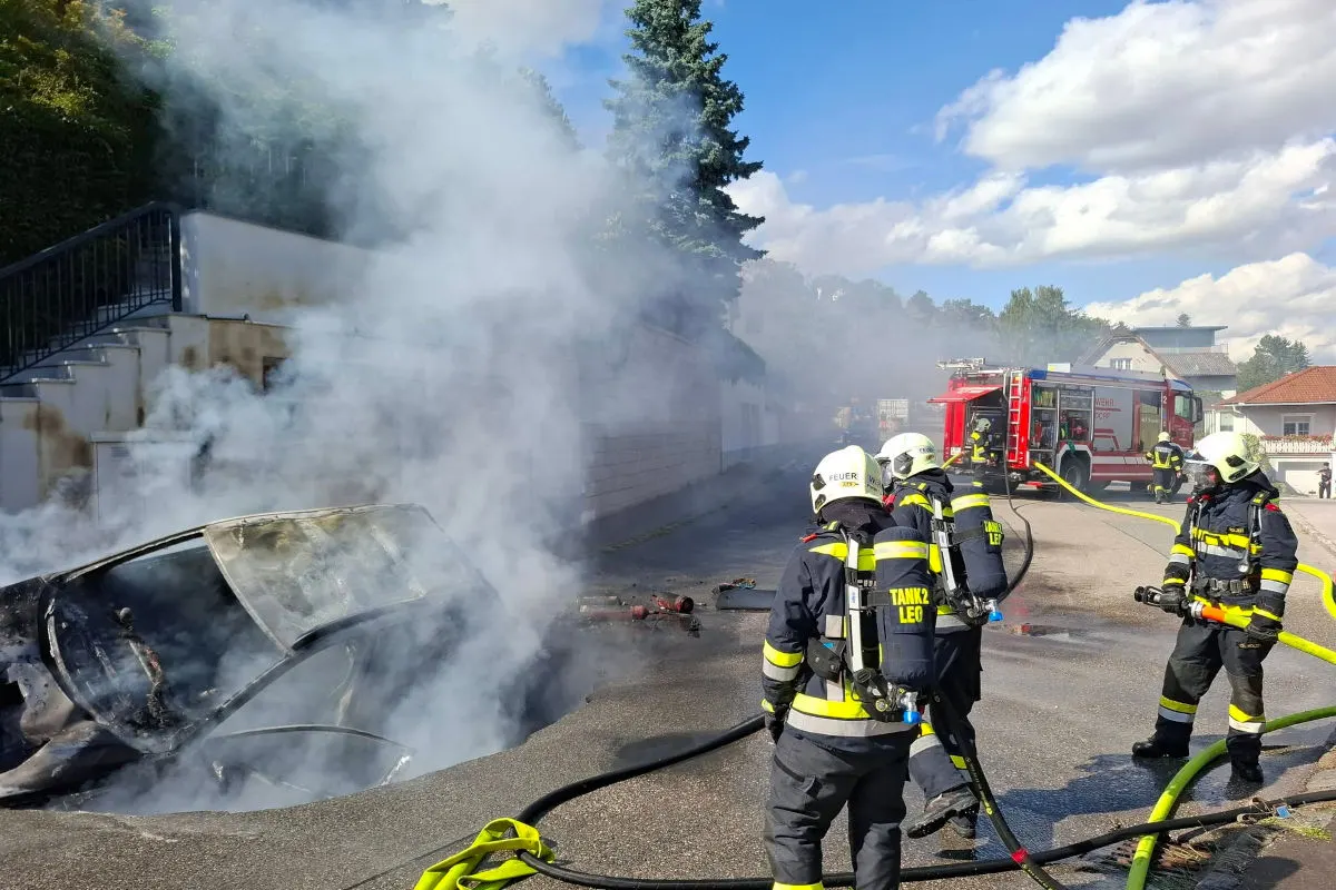 Mehrere Feuerwehrleute bekämpfen ein Feuer an einem stark rauchenden Auto. Im Hintergrund steht ein Feuerwehrfahrzeug in einem Wohngebiet.