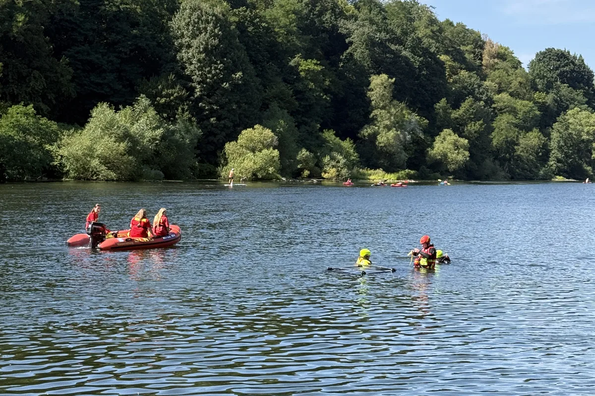 Ein Schlauchboot mit Rettungskräften und zwei Personen im Wasser. Im Hintergrund ist ein bewaldetes Ufer zu sehen.