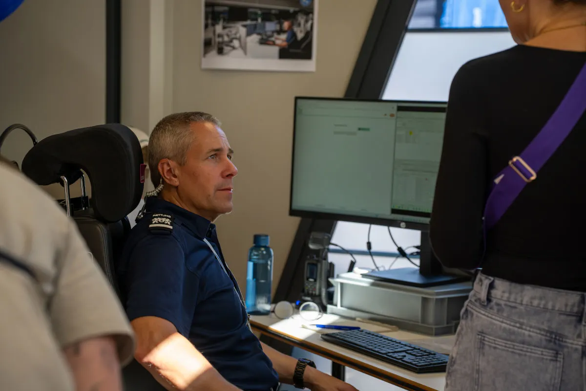 Ein Mann in einem blauen Rettungshemd sitzt an einem Schreibtisch vor einem Computerbildschirm. Eine Person steht vor ihm.