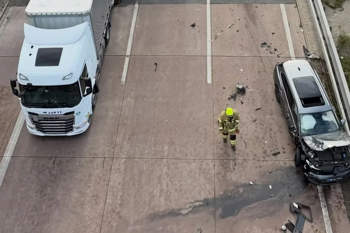 Ein Feuerwehrmann in gelber Schutzkleidung geht auf einer Straße zwischen einem großen weißen LKW und einem stark beschädigten silbernen Auto. Trümmerteile liegen verstreut.
