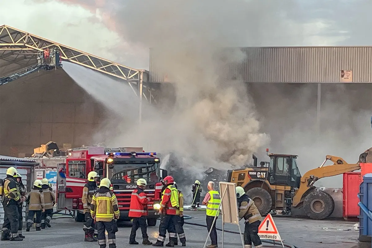 Mehrere Feuerwehrleute bekämpfen einen Brand in einer Industrieanlage. Ein Löschfahrzeug und schweres Gerät sind im Einsatz.