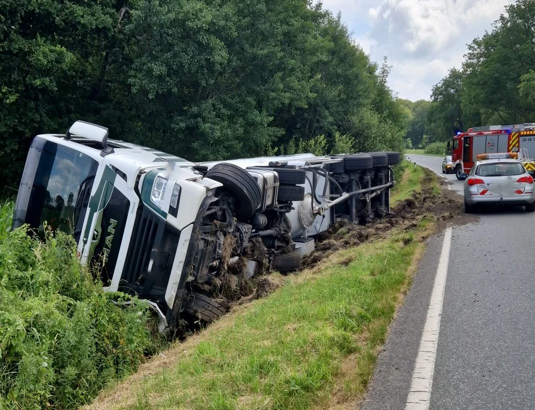 Ein schwerer Lkw liegt umgekippt in einem Straßengraben, während Einsatzfahrzeuge der Polizei und Feuerwehr an der Straße stehen.
