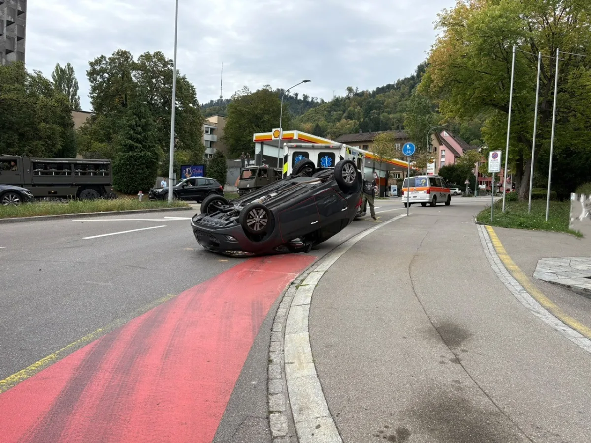 Ein dunkles Auto ist nach einem Unfall auf dem Dach liegend auf einer Straße zu sehen. Ein Polizeifahrzeug ist sichtbar, um die Unfallstelle abzusichern.