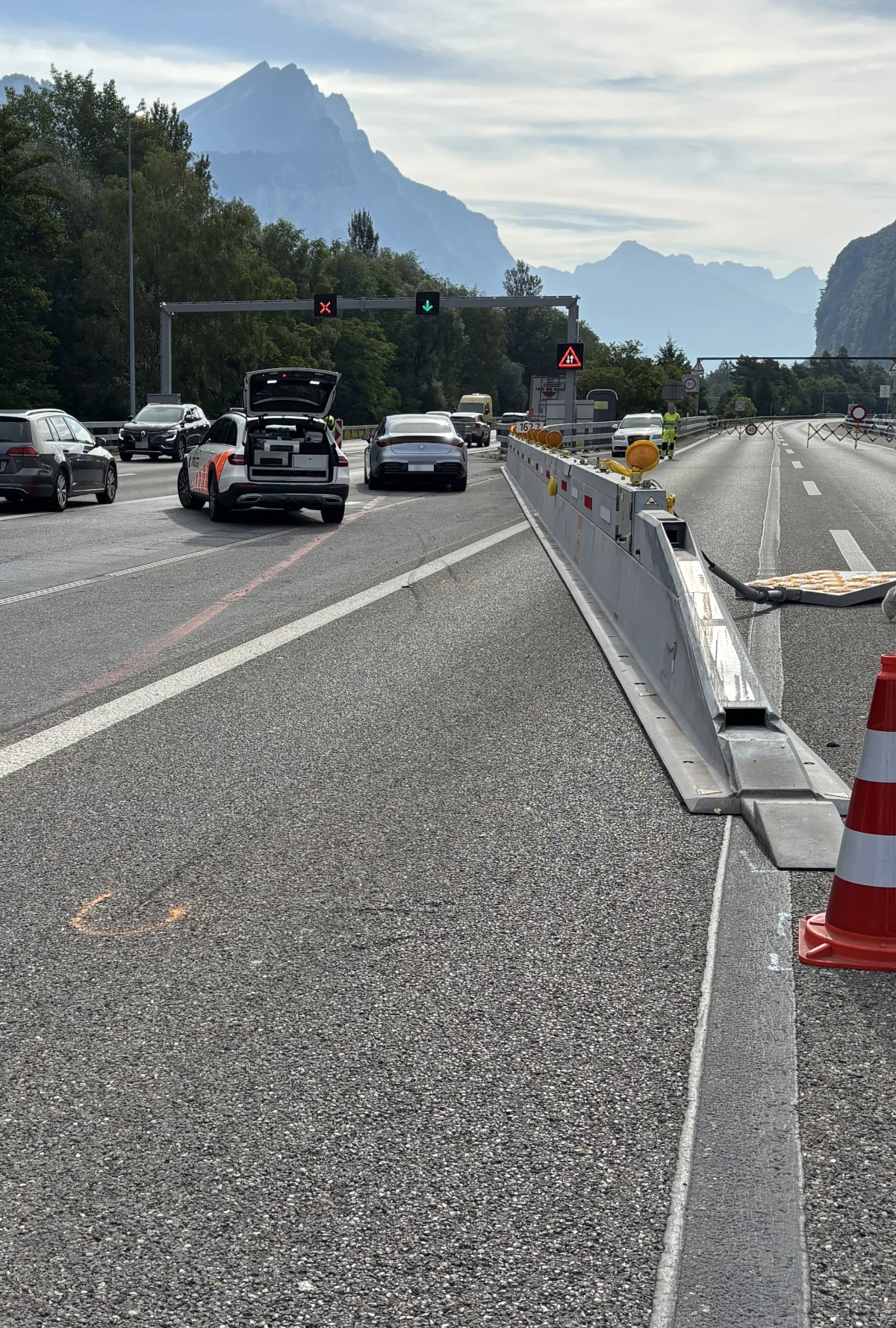 Ein Polizeifahrzeug steht auf einer Autobahn mit geöffnetem Kofferraum. Andere Autos sind auf der Fahrbahn sichtbar, und im Hintergrund erheben sich Berge. Absperrungen und Verkehrshinweise sind ebenfalls zu sehen.