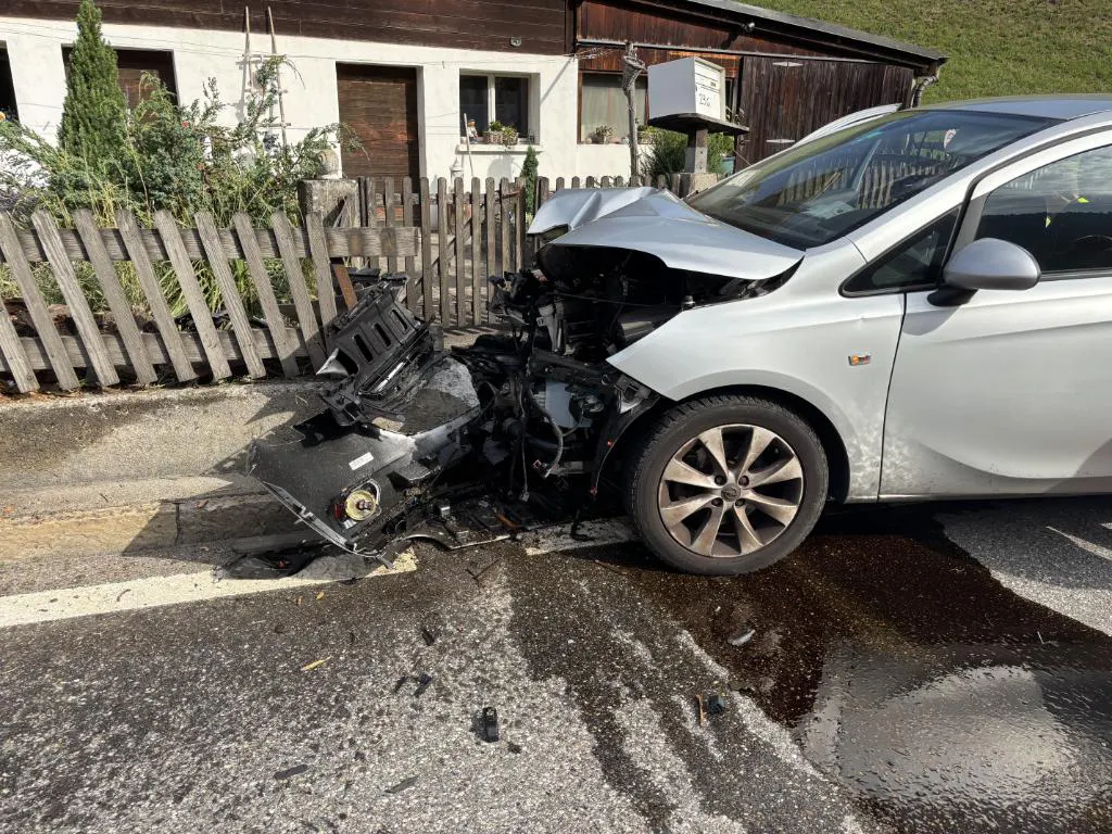 Ein silbernes Auto mit stark beschädigter Front steht vor einem Holzzaun in einem Wohngebiet. Der Asphalt ist nass, und Trümmer liegen verstreut auf der Straße.