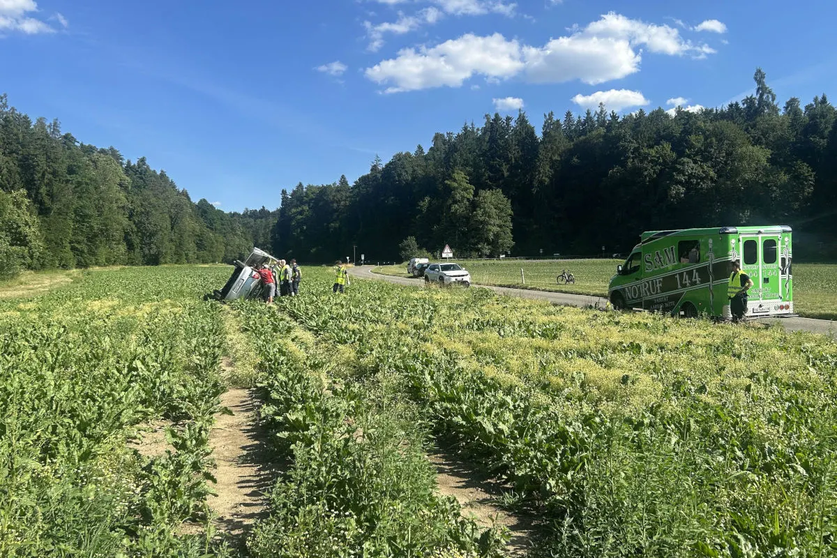 Das Bild zeigt ein Auto, das seitlich im Feld liegt. Mehrere Personen stehen herum, während ein grünes Einsatzfahrzeug in der Nähe auf der Straße parkt.