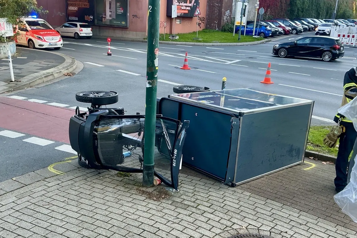 Ein umgekippter Anhänger liegt auf dem Gehweg, während im Hintergrund ein Polizeifahrzeug zu sehen ist. Verkehrskegel sichern die Stelle.