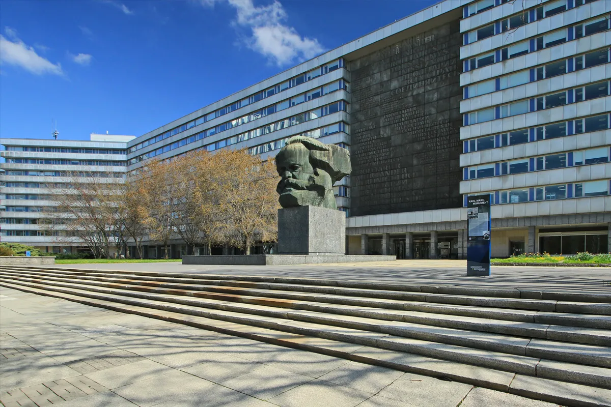Ein großes Karl-Marx-Monument steht zentral vor einem mehrstöckigen Gebäude. Der Himmel ist klar und blau, mit einigen Wolken.