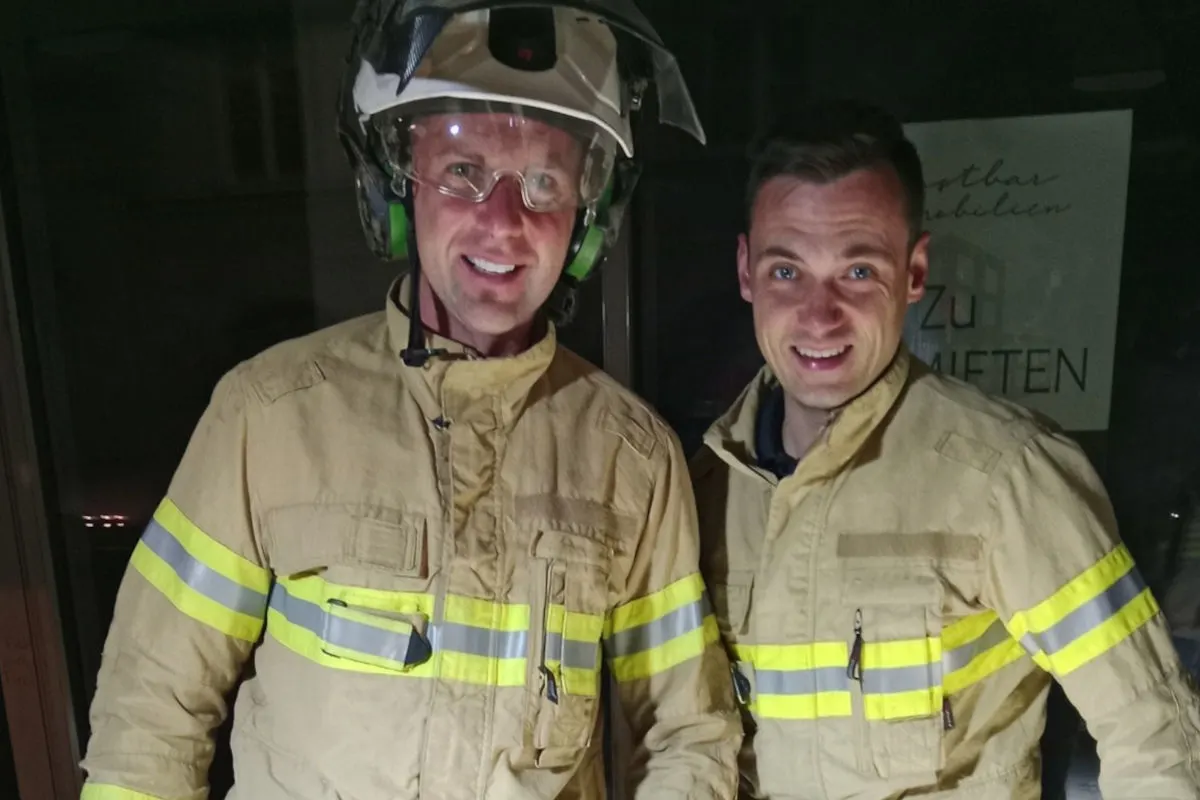 Zwei Feuerwehrmänner in beiger Schutzkleidung und Helmen stehen in einem Raum. Einer trägt einen Helm mit Visier hochgeklappt, beide lächeln.