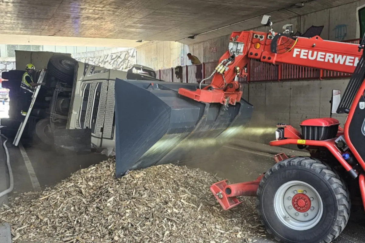 Ein Lastwagen liegt umgekippt in einer Unterführung, umgeben von verstreuten Holzschnitzeln und Feuerwehrgerät zur Bergung.