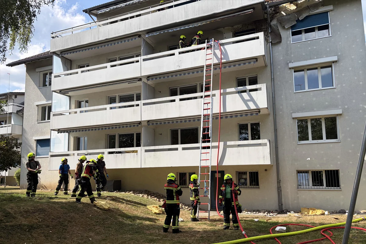 Eine Gruppe von Feuerwehrleuten arbeitet an einem vierstöckigen Wohngebäude. Sie benutzen eine Leiter, die an der Fassade lehnt.