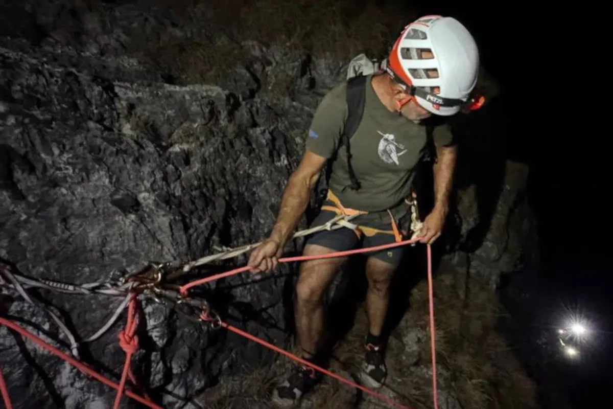 Ein Mann mit Schutzhelm und Sicherheitsgeschirr befestigt Kletterseile an einem Felsen in einer nächtlichen Szenerie.