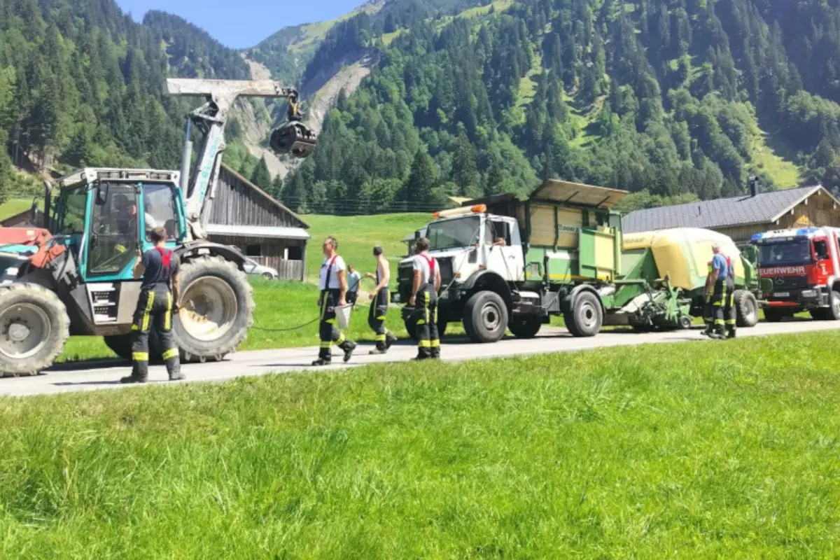 Eine Gruppe von Feuerwehrleuten steht neben einem Traktor und einem Feuerwehrfahrzeug in einer ländlichen Umgebung mit Bergen im Hintergrund.