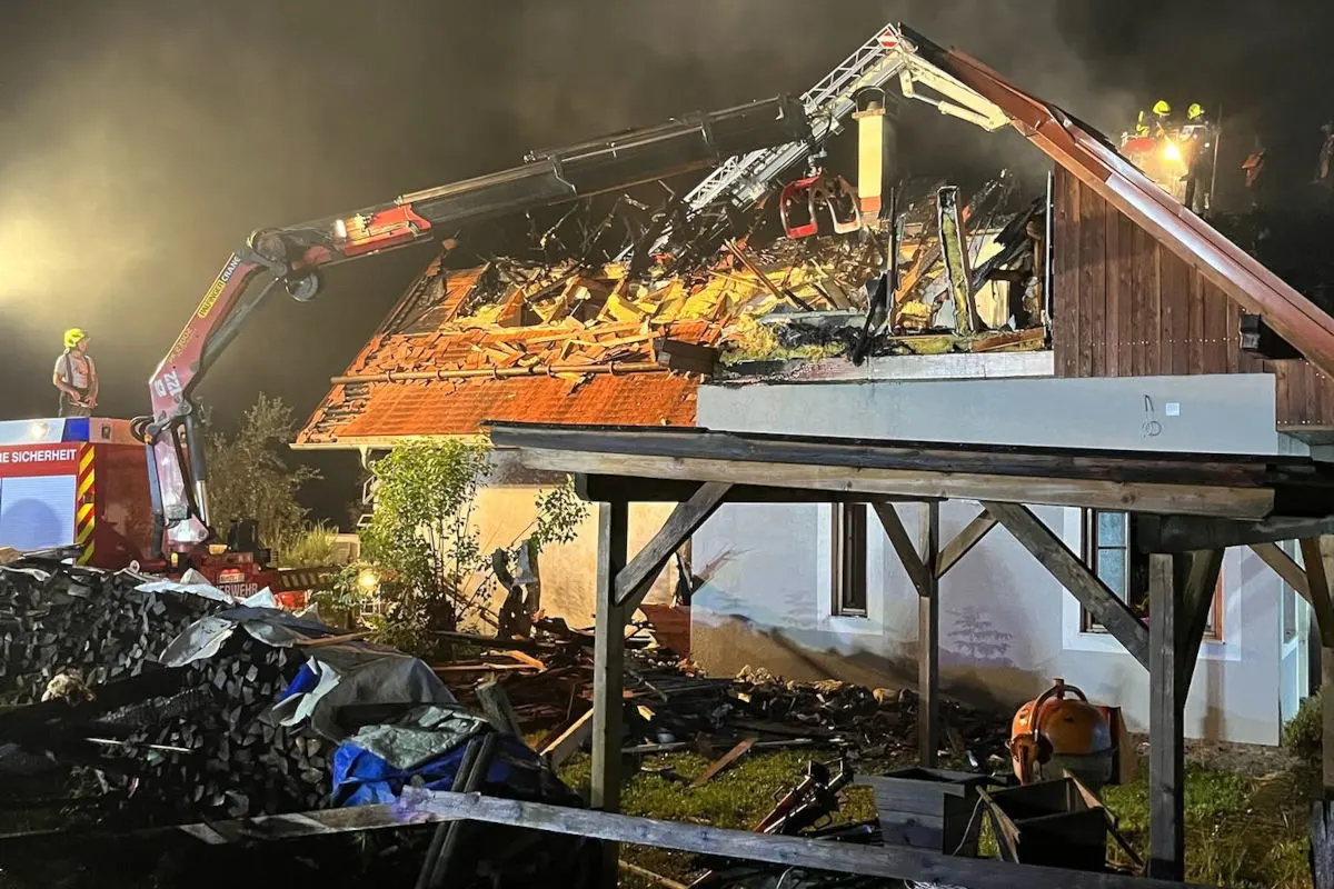 Ein Hausdach brennt in der Nacht, Feuerwehrleute setzen Hebebühne zur Brandbekämpfung ein. Beschädigungen sind deutlich sichtbar.
