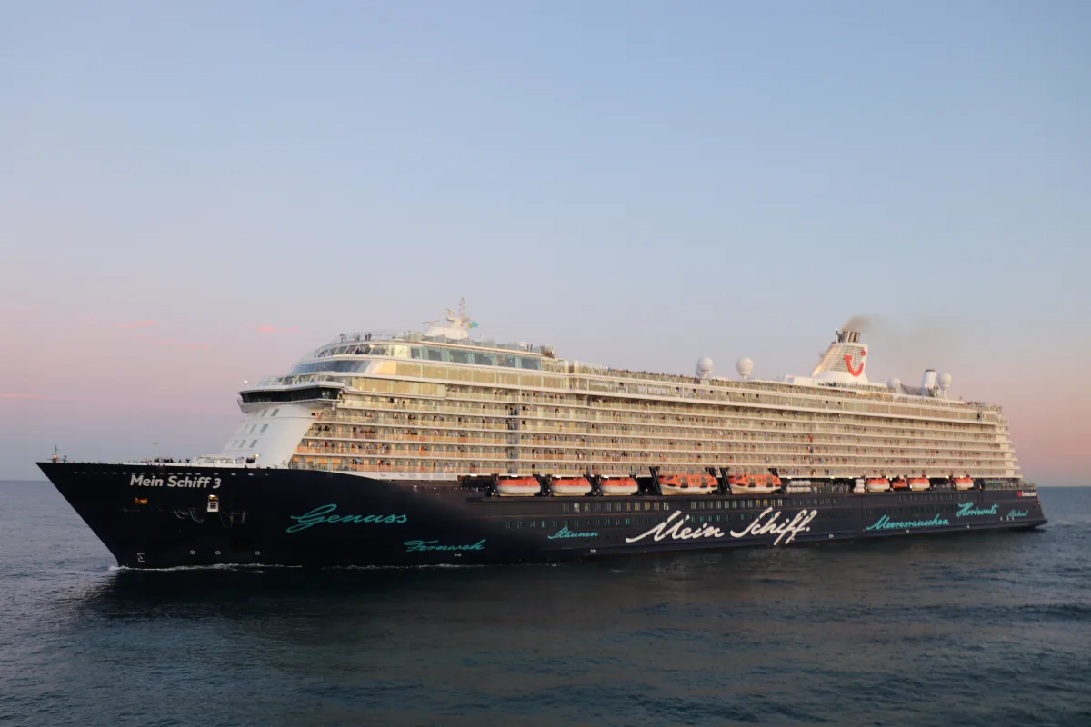 Ein großes Kreuzfahrtschiff mit dem Namen 'Mein Schiff 3' fährt auf dem Meer. Der Himmel zeigt einen sanften Sonnenuntergang.