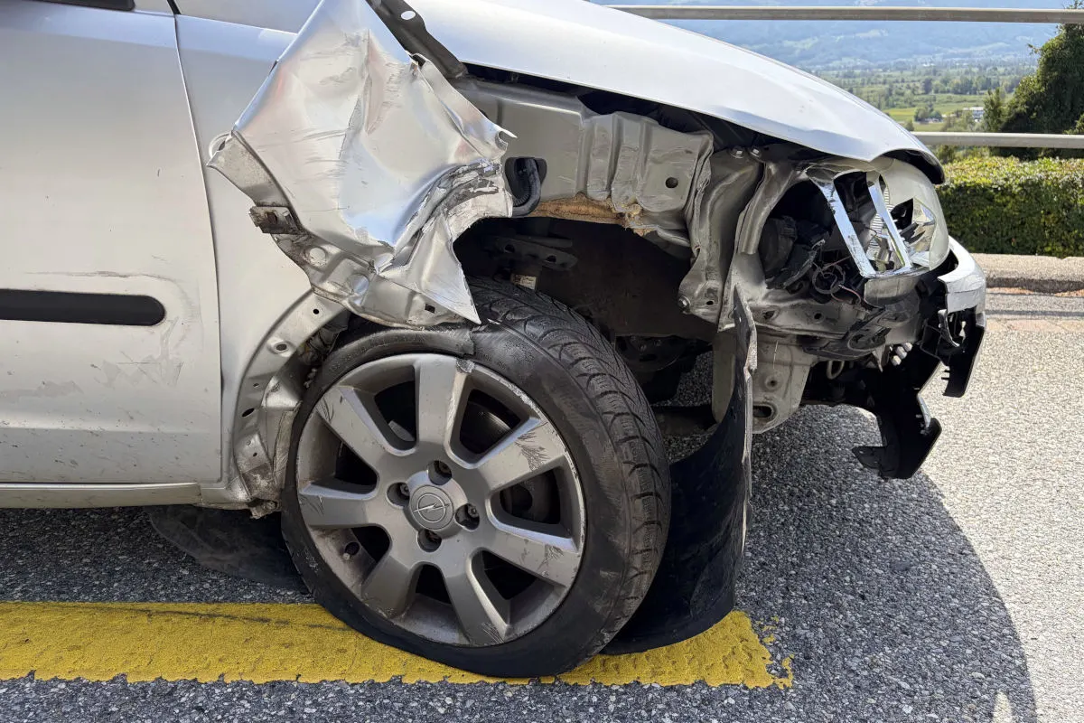 Das Bild zeigt die Nahaufnahme eines silbernen Autos mit einem stark verformten Kotflügel und zerstörter Frontpartie. Es steht auf einer Straße mit gelber Linie im Vordergrund.