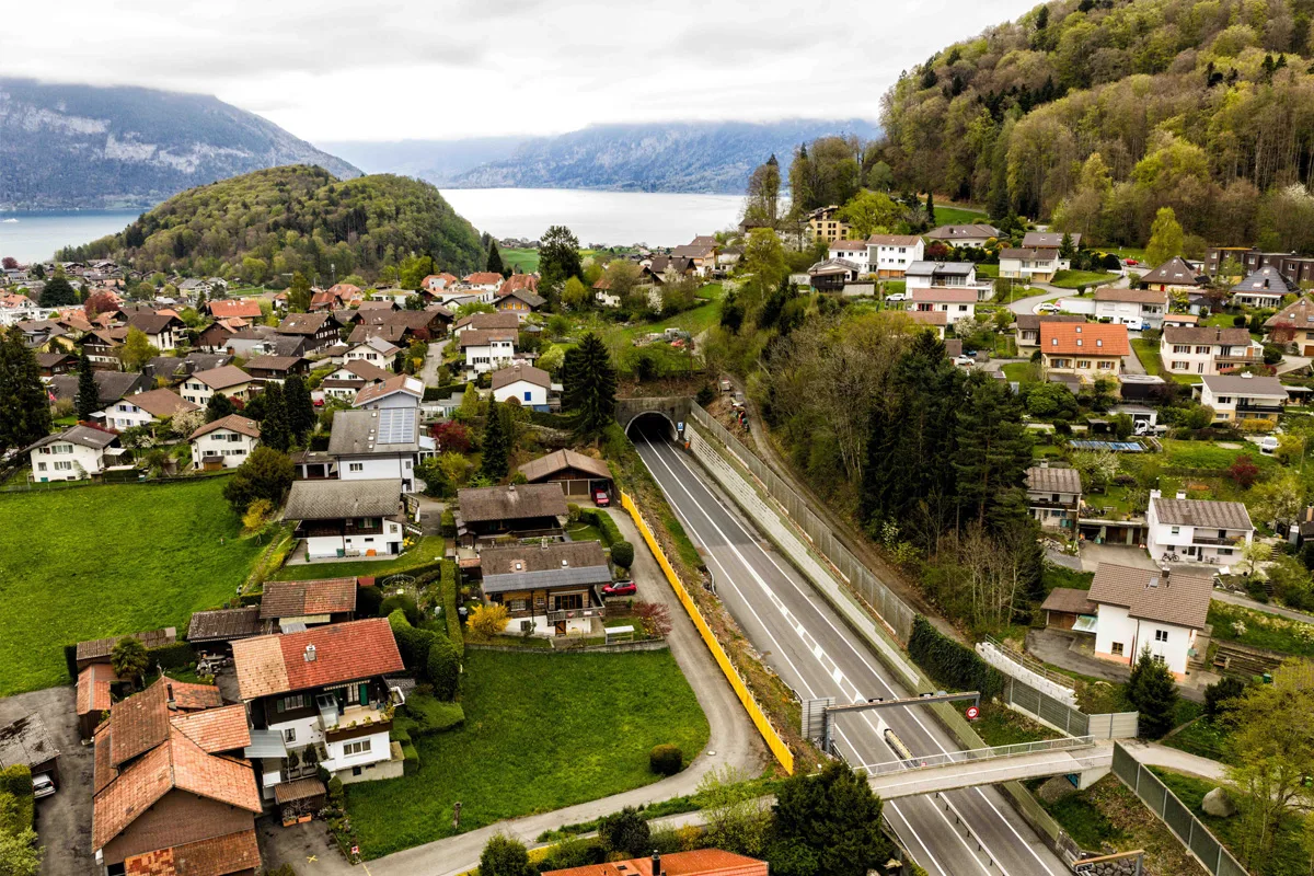Die Luftaufnahme zeigt einen Tunnel auf einer Straße, umgeben von Wohnhäusern, grünen Hügeln und einem See im Hintergrund. Die Vegetation ist üppig, und das Wetter wirkt bewölkt.