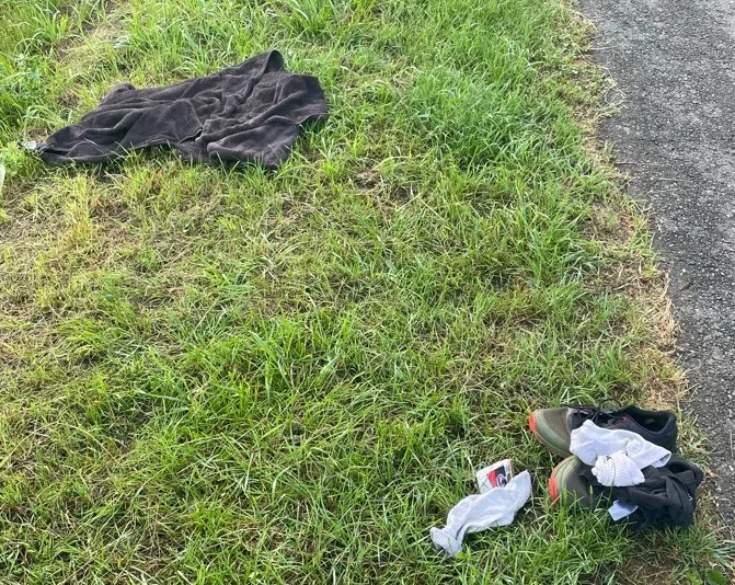 Ein dunkles Handtuch, ein Paar Schuhe und weiße Socken liegen auf einer Wiese neben einem Pfad.