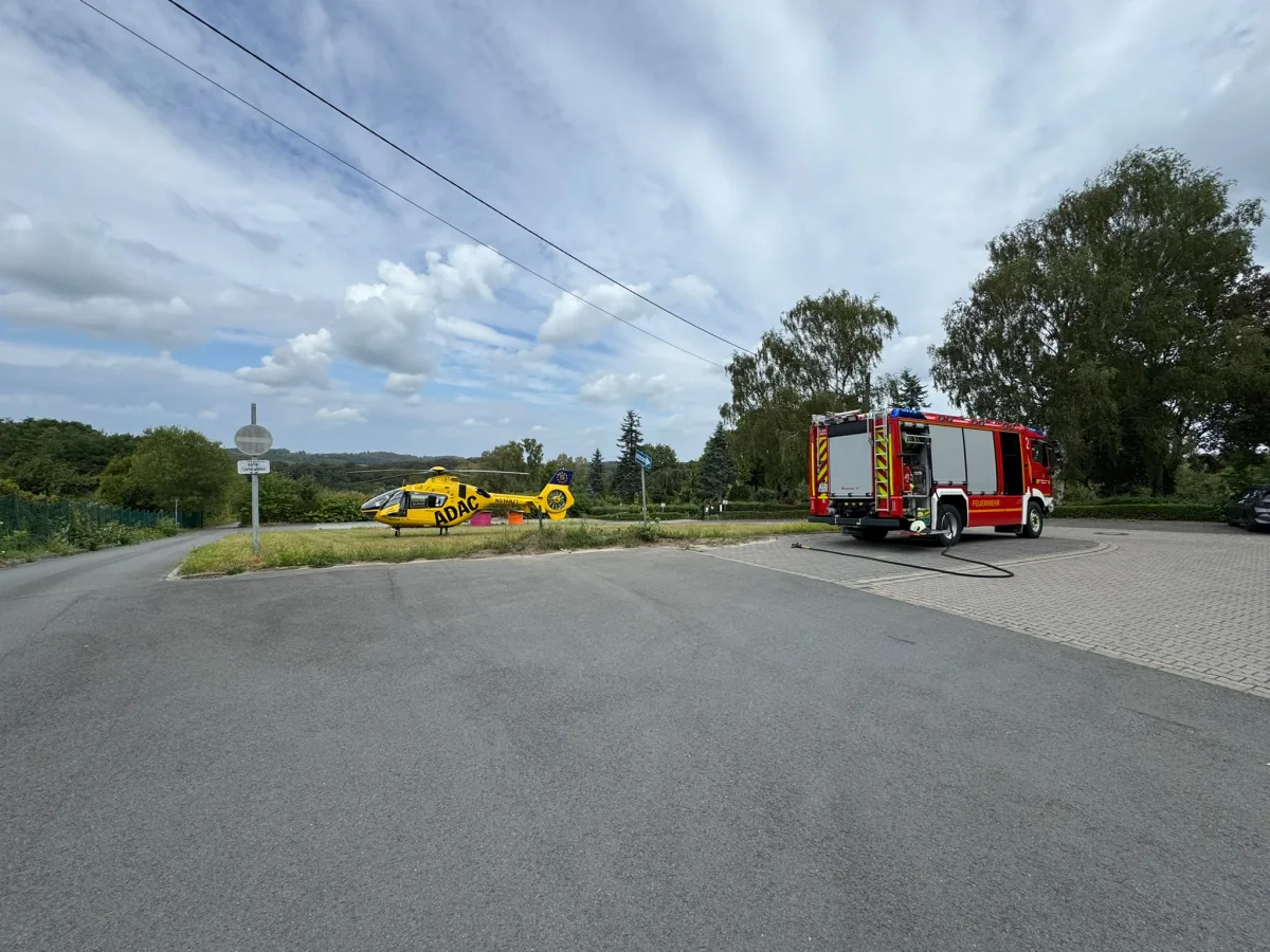 Ein gelber ADAC-Rettungshubschrauber und ein rotes Feuerwehrfahrzeug stehen auf einem ländlichen Parkplatz bei klarem Himmel.