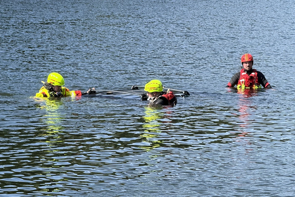 Drei Rettungskräfte in Tauchanzügen und Helmen stehen neben einem teilweise unter Wasser liegenden Fahrzeug.