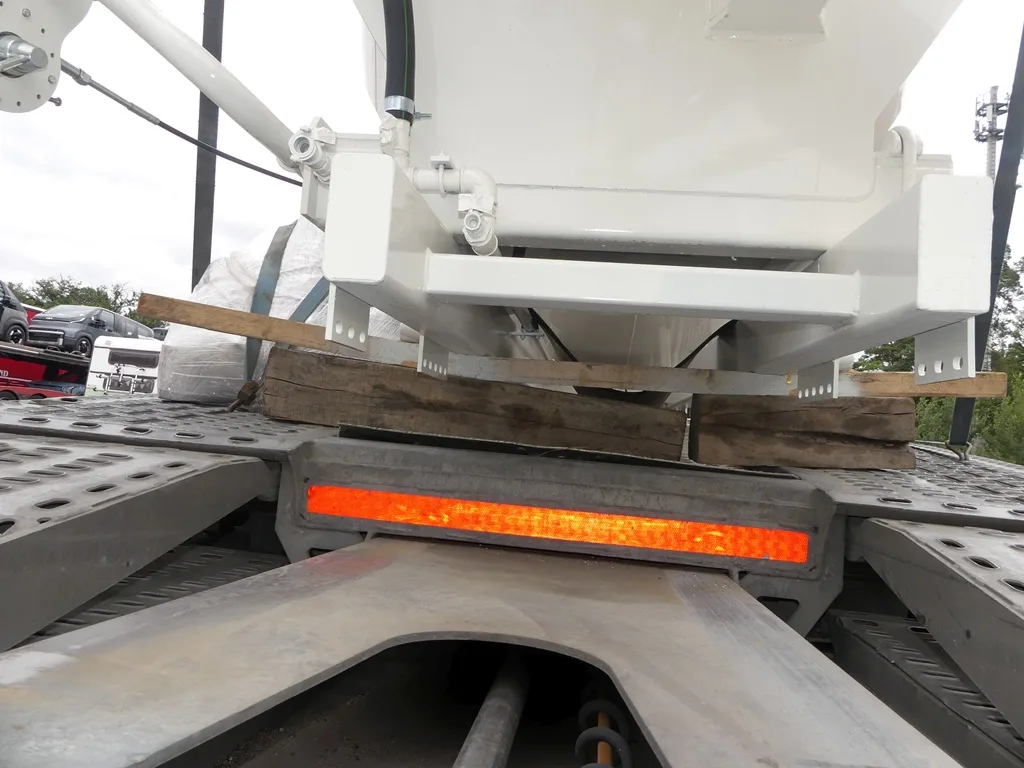 Ein weißer Behälter ist mit Holzplatten auf einem Lastwagen befestigt. Eine orangefarbene Warnmarkierung ist sichtbar.