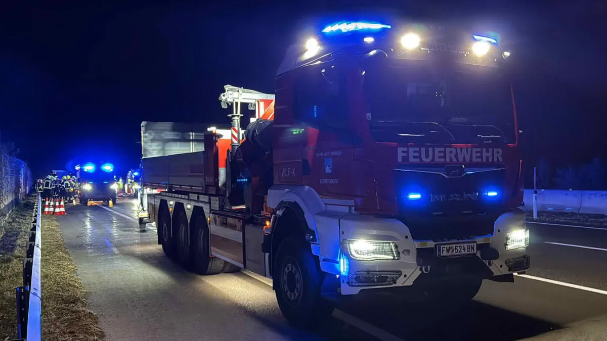 Ein großes rotes Feuerwehrauto steht mit eingeschaltetem Blaulicht auf einer nächtlichen Straße. Im Hintergrund sind weitere Einsatzfahrzeuge und Feuerwehrleute zu sehen.