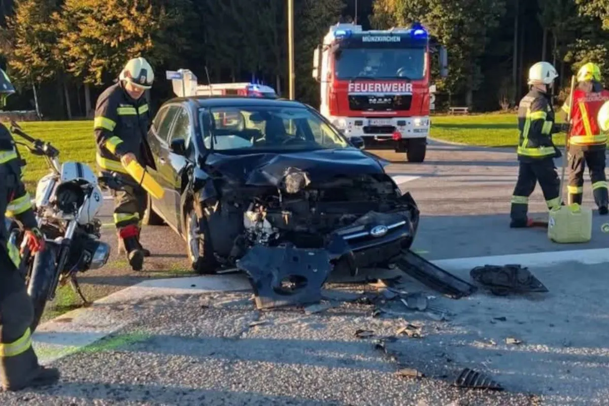 Ein Auto mit stark beschädigter Vorderseite steht auf der Straße. Feuerwehrleute sichern den Bereich ab, ein Feuerwehrfahrzeug ist im Hintergrund zu sehen.