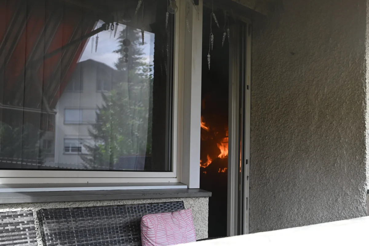 Ein Feuer brennt im Inneren eines Gebäudes, sichtbar durch eine geöffnete Terrassentür und Fenster mit teilweise geschlossenen Jalousien. Auf der Terrasse stehen Möbelteile.