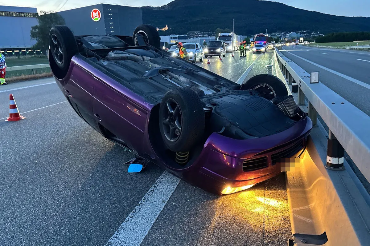 Ein violettes Auto liegt nach einem Unfall auf dem Dach auf einer Autobahn. Im Hintergrund sind Rettungskräfte und weitere Fahrzeuge zu sehen.