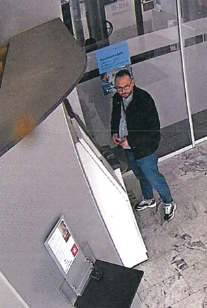 Ein Mann mittleren Alters steht vor einem Schalter in einem Gebäude, und schaut zur Kamera. Er trägt eine Jacke und Jeans.