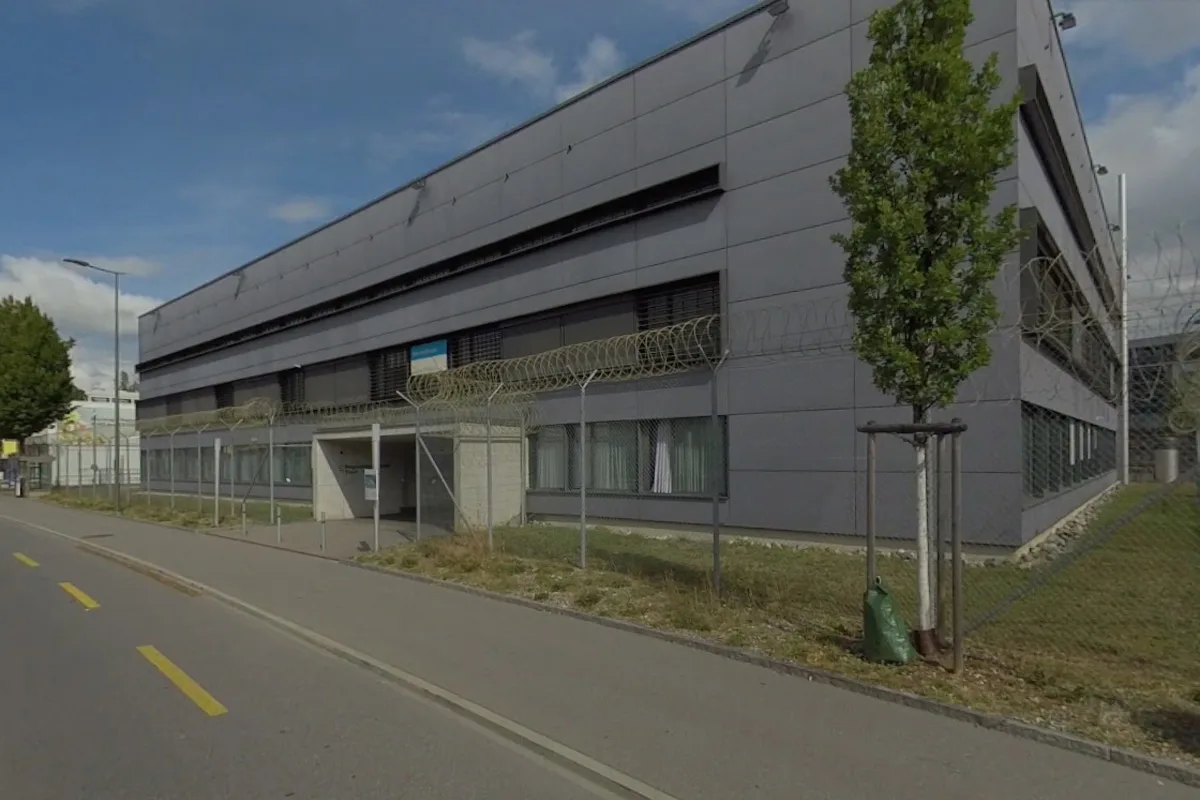 Das Bild zeigt ein modernes, graues Gebäude unter blauem Himmel, geschützt durch einen Stacheldrahtzaun an der Vorderseite. Ein Gehweg verläuft entlang der Straße vor dem Gebäude.