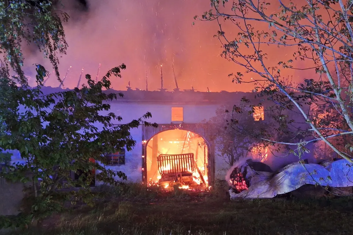 Ein Gebäude steht in Flammen. Das Dach brennt stark, während Funken in den Nachthimmel aufsteigen. Bäume rahmen das Bild ein.