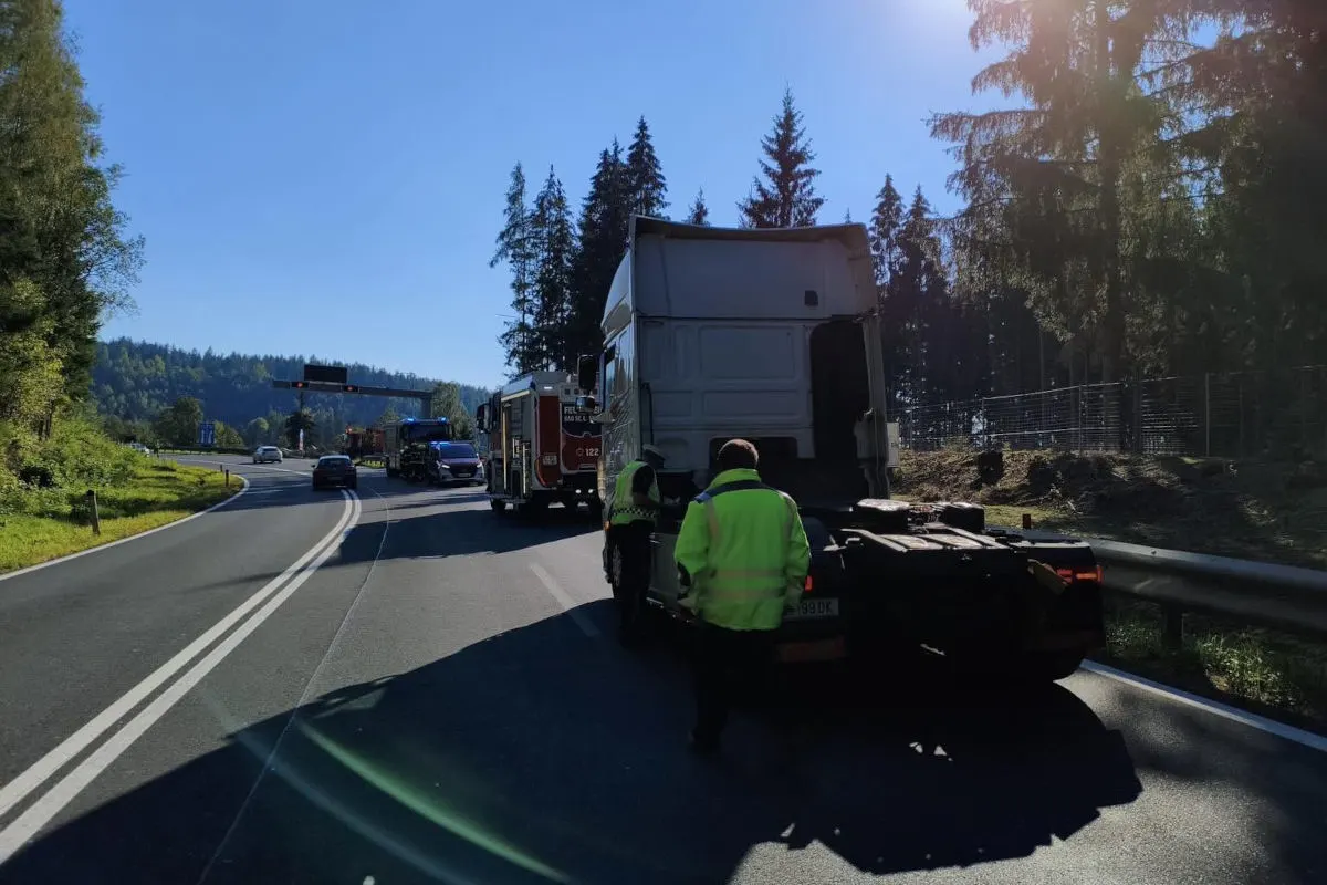 Ein Lkw steht auf einer sonnigen Landstraße, begleitet von Feuerwehrfahrzeugen. Zwei Personen in Warnwesten sind sichtbar.