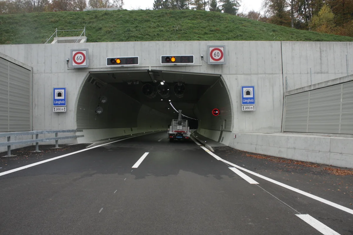 Ein Fahrzeug fährt in einen beleuchteten Straßentunnel ein. An der Wand steht 'Längholz', mit einer Längenangabe von 2490 Metern.