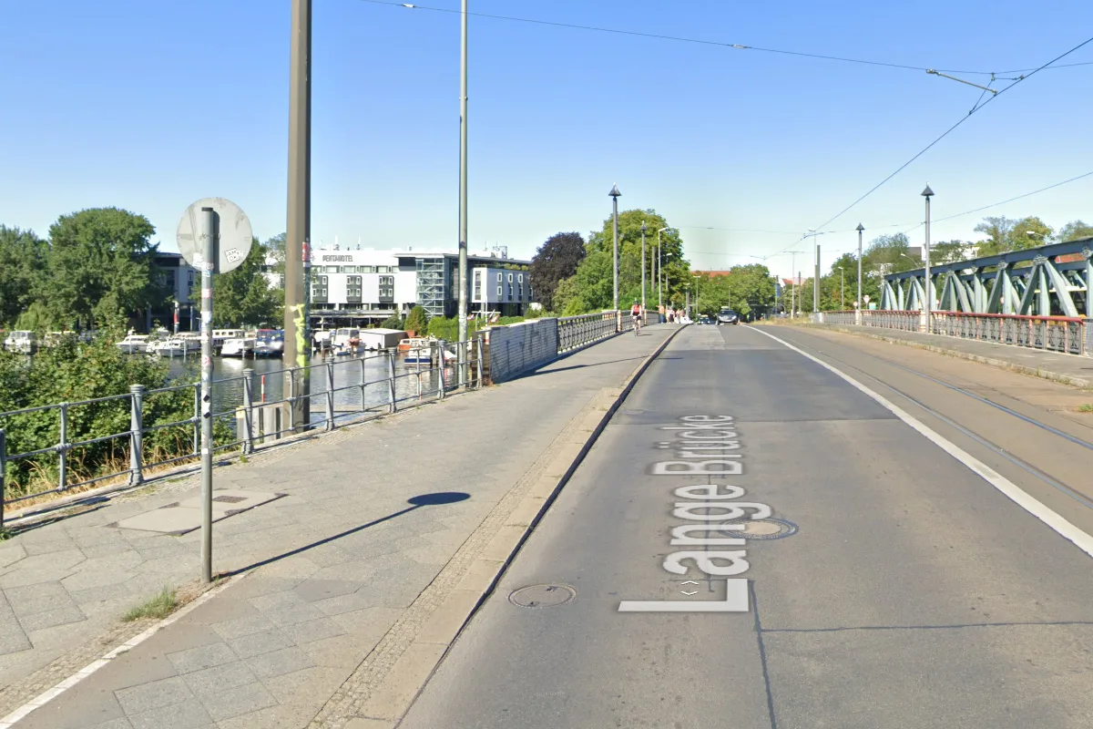 Eine Brücke mit Straßen- und Fußgängerbereich neben einem Fluss bei klarem Himmel. Vereinzelt sind Fußgänger sichtbar.