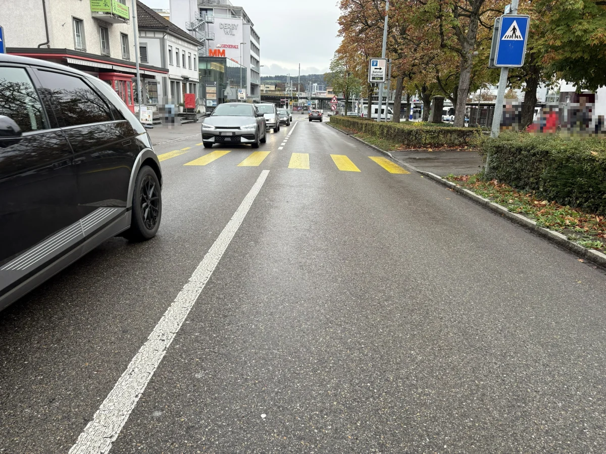 Mehrere Fahrzeuge fahren auf einer städtischen Straße vorbei an einem Zebrastreifen. Die Blätter an den Bäumen sind herbstlich gefärbt.
