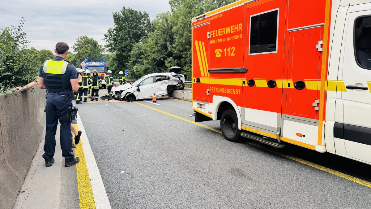 Ein beschädigtes weißes Auto steht auf der Autobahn neben einem Feuerwehrfahrzeug. Polizisten und Feuerwehrleute sind vor Ort.