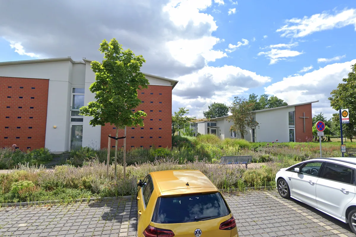Im Vordergrund steht ein gelbes Auto auf einem Parkplatz. Im Hintergrund sind moderne Gebäude mit Kreuz und Ziegelfassade zu sehen.