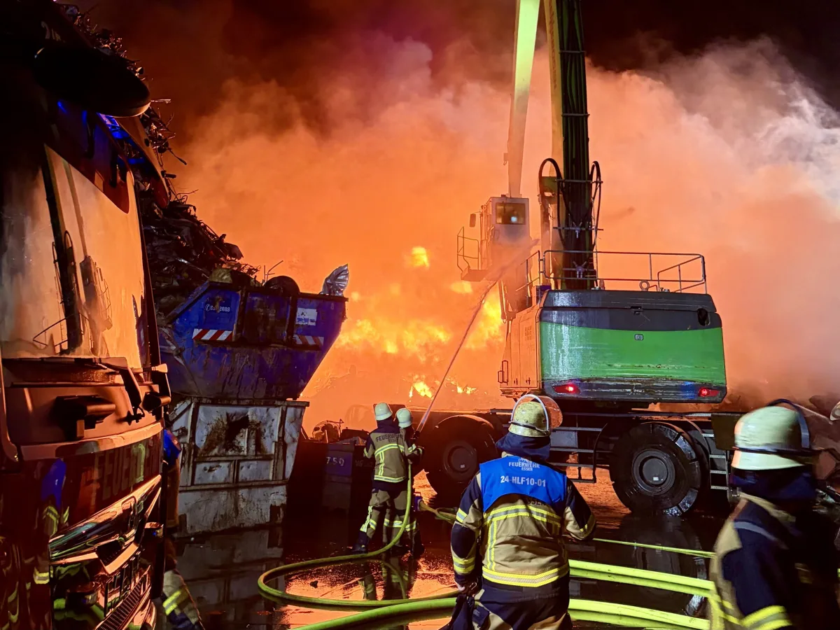 Mehrere Feuerwehrleute sind im Einsatz, um ein intensives Feuer auf einem Schrottplatz zu löschen. Ein großer Bagger betätigt einen Löschschlauch, während Flammen und Rauch den Hintergrund dominieren.