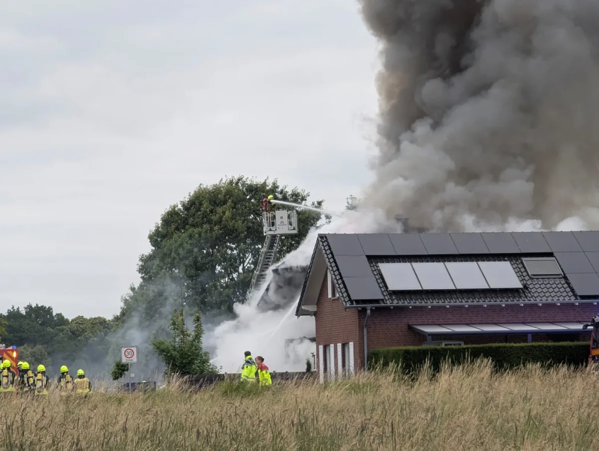 Rauch steigt von einem Wohnhaus auf, Feuerwehrleute in Schutzkleidung bekämpfen das Feuer mit einem Schlauch von einer Leiter aus.
