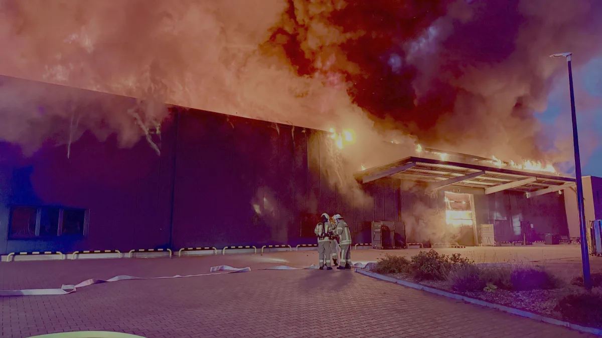 Dichte Rauchwolken und Flammen steigen aus einem Gebäude auf, während zwei Feuerwehrleute darunter stehen.