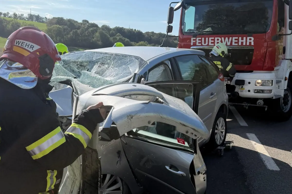 Ein Feuerwehrmann in voller Ausrüstung steht neben einem stark beschädigten silbernen Auto. Im Hintergrund ist ein rotes Feuerwehrfahrzeug zu sehen.